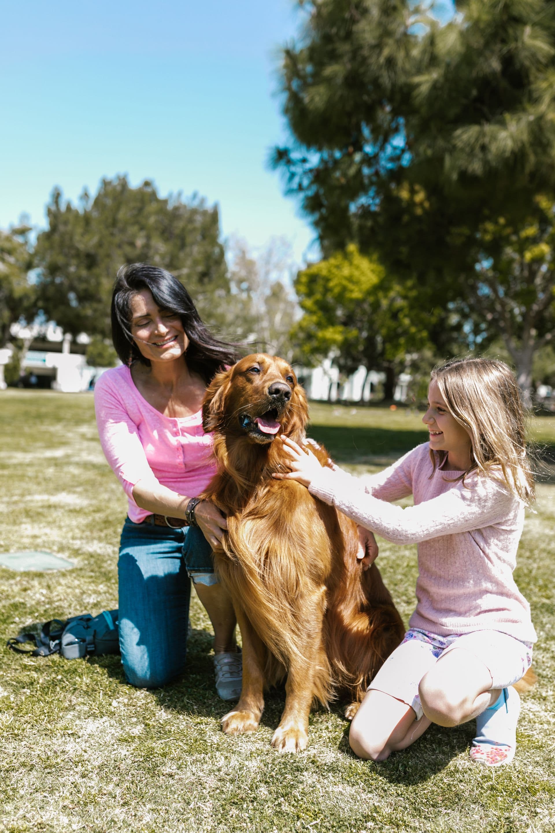 A mother and daughter laughing with their golden retriever in a park